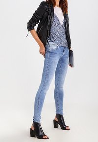 Chaqueta de cuero negra sobre una blusa de patrón azul, jeans skinny de un azul claro y botas de tacón bloque abierto en negro. Bolso de mano en la mano.