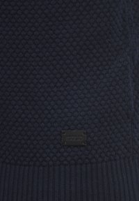 Marineblauwe gebreide trui met een gestructureerd honingraatpatroon, voorzien van een geribbelde zoom en een zwart label met "USOLID" aan de onderkant.