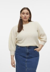 Maglione lavorato a maglia color crema con maniche a sbuffo, abbinato a una gonna a tubino in denim. La modella ha i capelli castani di lunghezza spalla e indossa orecchini pendenti.