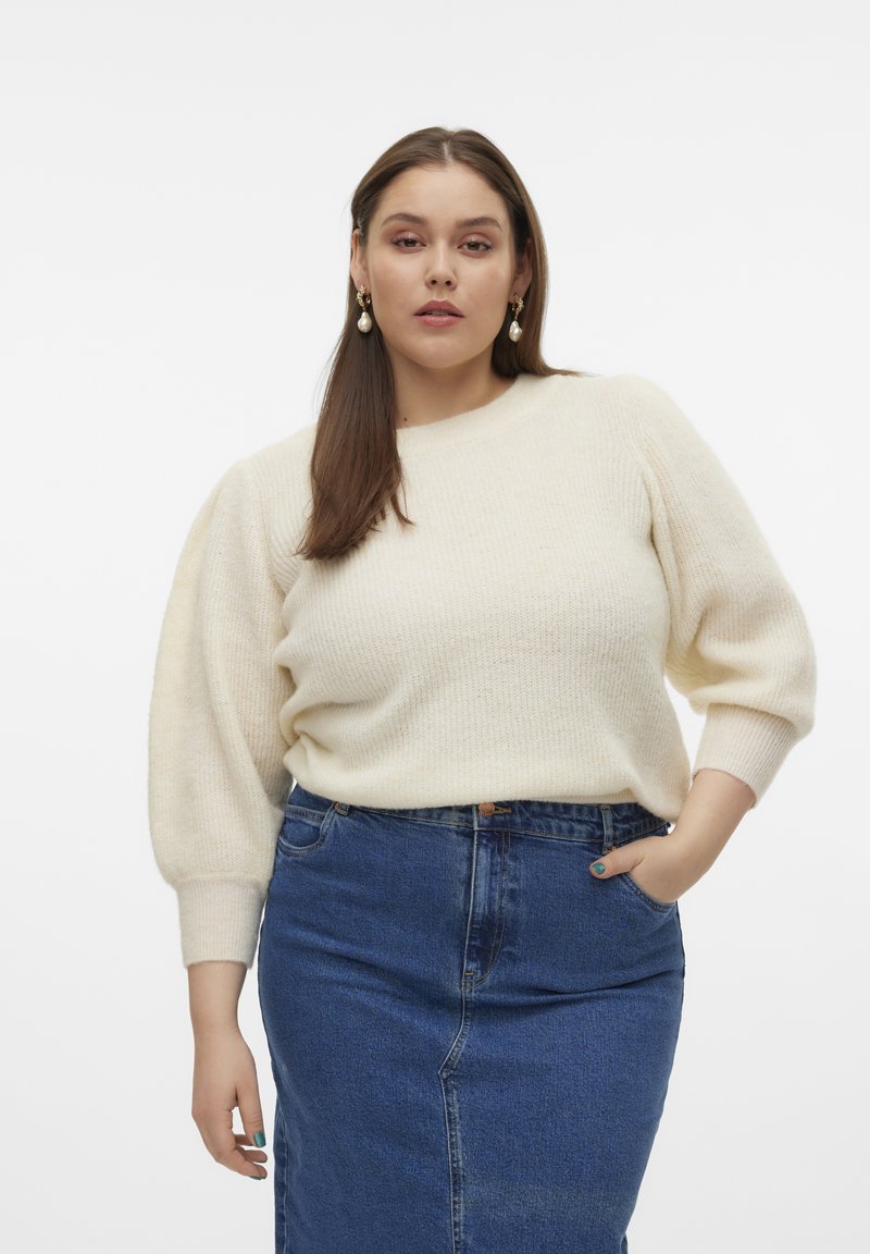 Maglione lavorato a maglia color crema con maniche a sbuffo, abbinato a una gonna a tubino in denim. La modella ha i capelli castani di lunghezza spalla e indossa orecchini pendenti.
