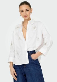 Camicia bianca con bottoni, decorata con brillantini sul davanti, abbinata a jeans a gamba larga blu scuro. Tessuto liscio, vestibilità comoda.