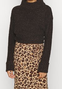 Dunkelbrauner, grob gestrickter Pullover mit hohem Stehkragen und lockerer Passform, kombiniert mit einem beigen Rock mit Leopardenmuster und Bindegurt.