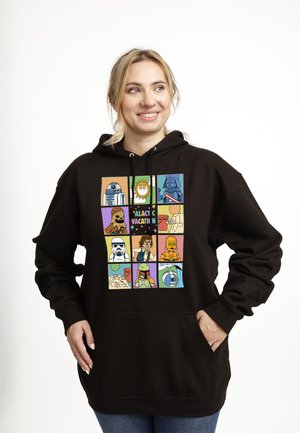 Kvinde smilende, iført sort hættetrøje med farverige tegneserie-agtige Star Wars-figurer i gitterpaneler og teksten "Galaktisk ferie".