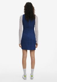 Rochie din denim albastru cu un corset ajustat și fustă evazată, având un model texturat. Mâneci lungi, transparente, cu dungi sunt vizibile dedesubt.