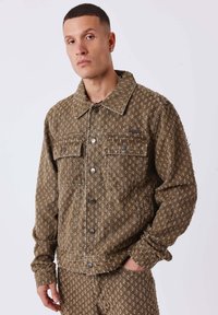 Homme portant un ensemble coordonné de veste et pantalon brun texturé avec de petits motifs clairs, posant avec une main dans la poche contre un fond blanc.