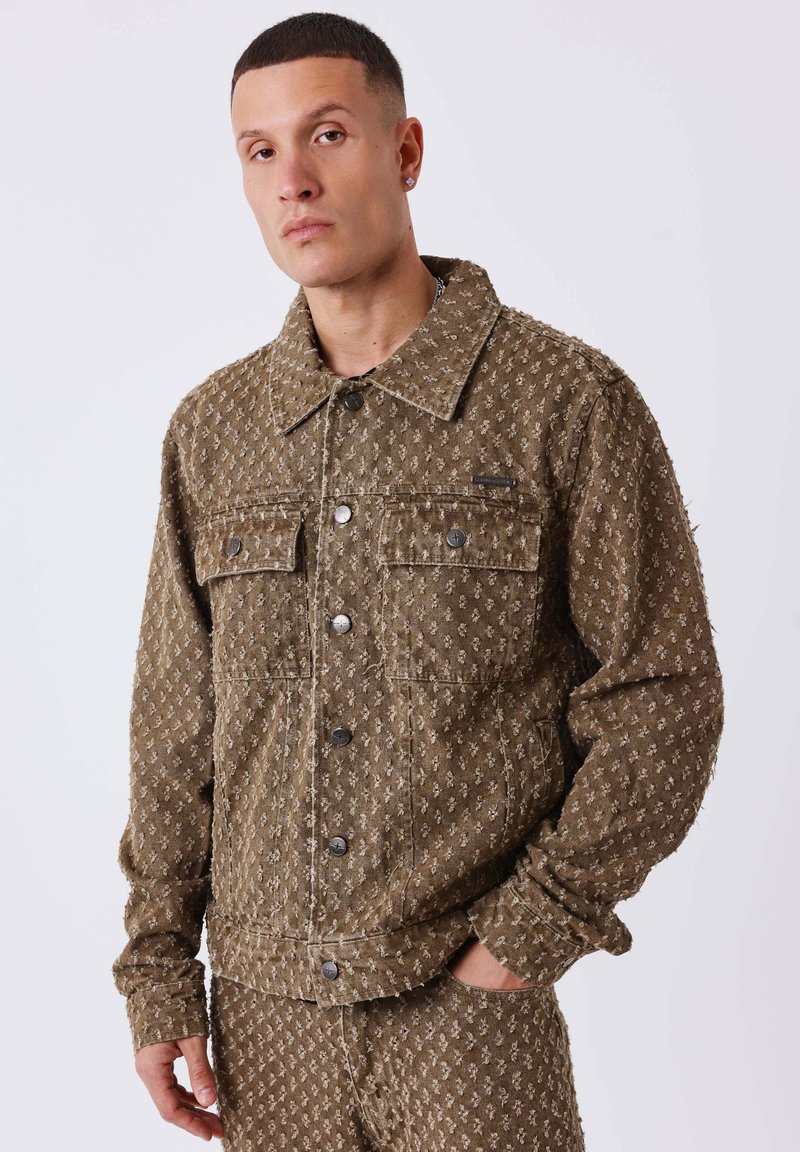 Homme portant un ensemble coordonné de veste et pantalon brun texturé avec de petits motifs clairs, posant avec une main dans la poche contre un fond blanc.