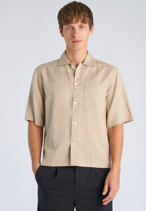 Młody mężczyzna ubrany w beżową, wzorzystą koszulę na krótki rękaw zapinaną na guziki oraz czarne spodnie, stojący z rękami w kieszeniach na jasnym tle.