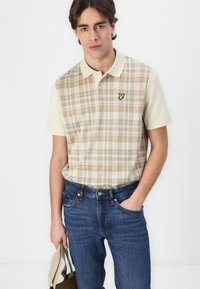 Camisa polo con cuello en un patrón de cuadros beige, con acentos naranjas. Combinada con jeans azules y una bolsa de mano verde, diseño minimalista.