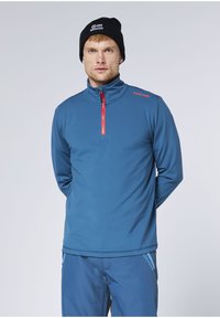 Mann trägt eine blaue Jacke mit Reißverschluss und passende Hose mit dem "Chiemsee"-Logo sowie eine schwarze Mütze mit einem Plus-Minus-Symbol.