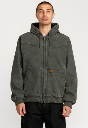 Homme aux cheveux courts portant un sweat à capuche gris foncé zippé avec capuche et poches avant, debout devant un fond clair uni.