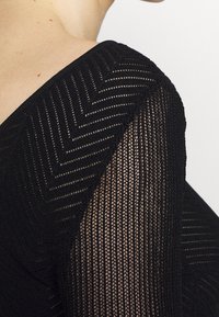 Czarny sweter z teksturowanym dzianinowym materiałem w wzór chevron na rękawie, prezentujący półprzezroczysty wygląd.