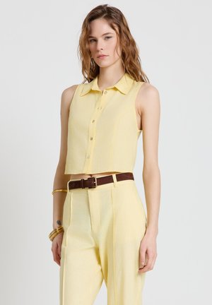 Jeune femme aux cheveux ondulés portant un crop top jaune sans manches à boutons et un pantalon jaune taille haute avec ceinture marron et bracelets dorés.