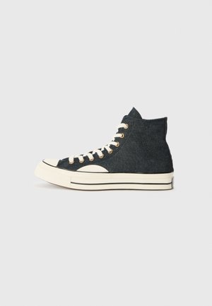 Zapatilla alta de mezclilla negra con suela de goma blanca, puntera blanca, cordones blancos y ojales dorados, mostrada de perfil sobre un fondo claro.