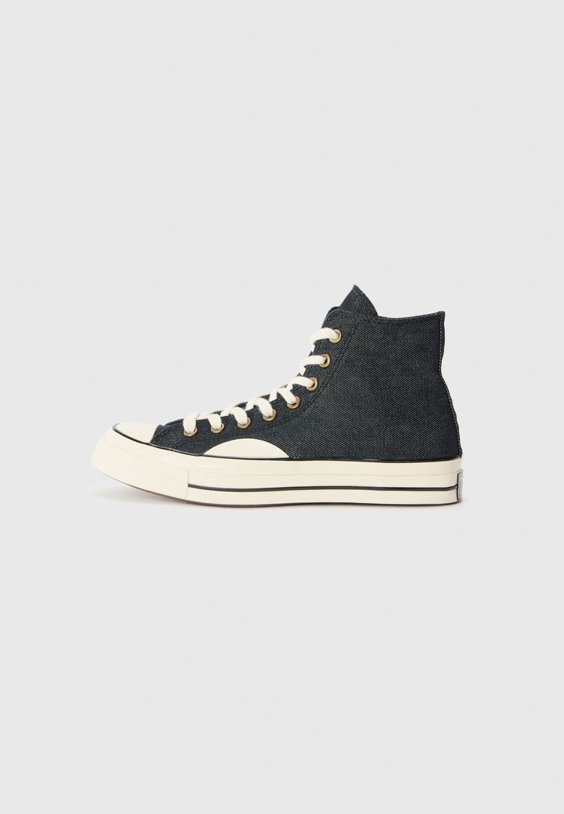 Baskets montantes en denim noir avec semelle en caoutchouc blanche, bout blanc, lacets blancs et œillets dorés, vues de profil sur un fond clair.