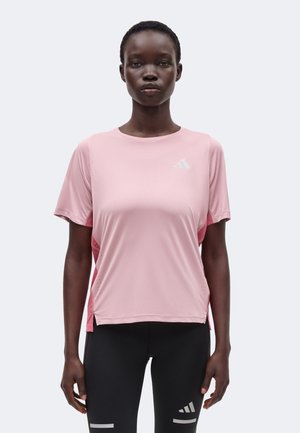 Femme portant un t-shirt de sport rose clair à manches courtes et des leggings noirs avec un logo, debout face à l'avant sur un fond uni.
