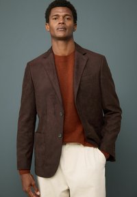 Hnědý semišový blazer se dvěma knoflíky, špičatými klopami a předními kapsami, vrstvený přes oranžový pletený svetr a krémové kalhoty.