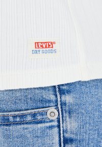 Tejido blanco con superficie acanalada con etiqueta de "Levi's Dry Goods", combinado con denim azul claro que presenta un botón de metal visible y detalles de costura.