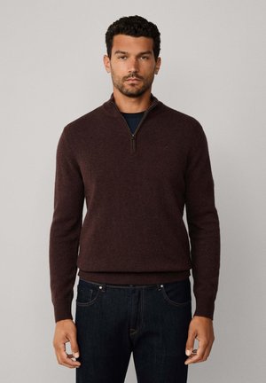 Brauner Pullover mit Reißverschluss am Hals, langen Ärmeln, gerippten Bündchen und Saum, aus einem weichen Material, kombiniert mit dunkelblauen Jeans.