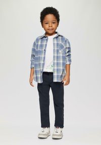 Blau-weiß kariertes Hemd über einem weißen T-Shirt, kombiniert mit marineblauen Hosen und weißen Sneakers mit grauen Akzenten.