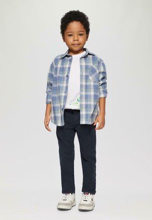 Chemise à carreaux bleue et blanche avec des manches boutonnées sur un T-shirt blanc, assortie à un pantalon marine et des baskets blanches avec des accents gris.