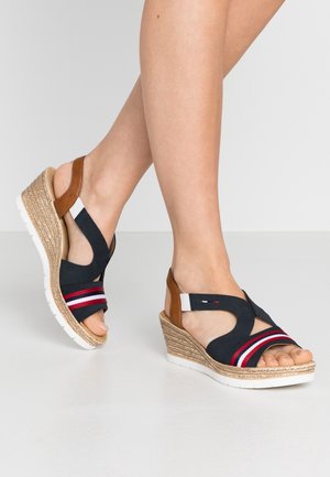 Benen in marineblauwe wedgesandalen met rode, witte en bruine banden, die over een gladde lichtgekleurde vloer lopen tegen een effen achtergrond.