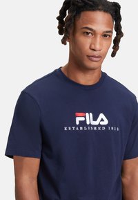 Camiseta de algodón navy con mangas cortas. Presenta un logo bordado de FILA en blanco y rojo y el texto "ESTABLECIDO EN 1911" en el pecho.