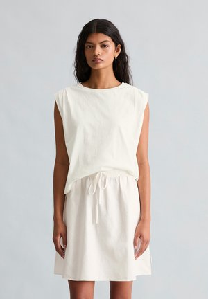 Kobieta z długimi ciemnymi włosami, ubrana w białą, bezrękawnikową bluzkę i pasującą spódnicę ze sznurkiem, stojąca na tle jednolitego jasnoszarego tła.