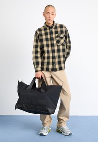 Black padded tote bag with dual handles, featuring a textured finish and logo patch. Model wears a tan and black checkered shirt and beige pants.