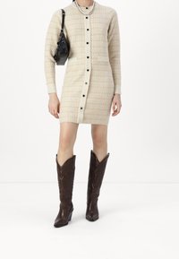 Robe en tricot beige avec un motif texturé, fermeture à boutons et deux poches. Associée à des bottes de cowboy en cuir marron avec des détails embossés.