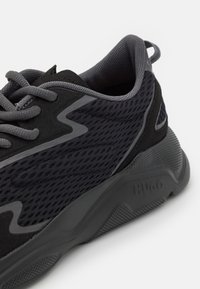 Scarpa sportiva nera e grigia con tomaia in rete, suola in gomma e dettagli testurizzati. Il design include un logo sul lato e materiali sovrapposti.