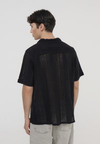 Maglietta nera a maniche corte con un design leggero, texturizzato e lavorato a maglia con un motivo aperto. Dettaglio del colletto posteriore visibile.
