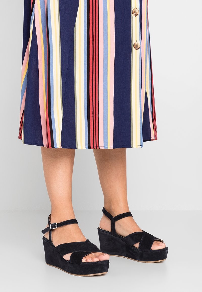 Jambes portant des sandales compensées en daim noir avec brides à la cheville, associées à une jupe mi-longue à rayures verticales multicolores.