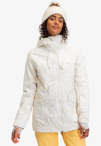Witte geïsoleerde jas met een gestructureerd patroon, een capuchon, rits- en zakken met een flap, gecombineerd met gele broek en een crèmekleurige gebreide muts.