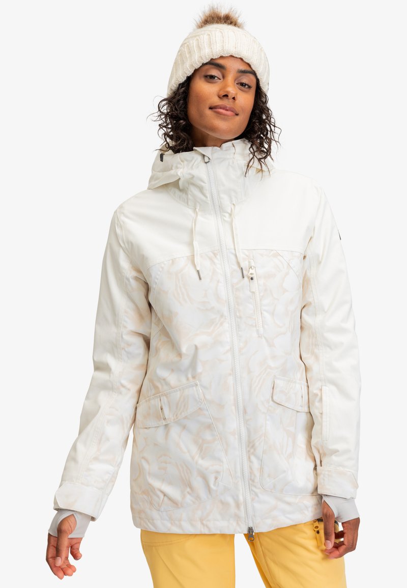 Witte geïsoleerde jas met een gestructureerd patroon, een capuchon, rits- en zakken met een flap, gecombineerd met gele broek en een crèmekleurige gebreide muts.