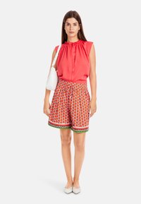 Red sleeveless top with ruffle neckline paired with patterned shorts featuring orange drop shapes, white accents, and a green trim. White handbag.