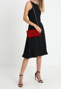 Musta hihaton mekko, jossa levenevä helma. Punainen mokkanahkainen clutch-laukku, jossa taitettava muotoilu, hopeinen ketjuhihna ja koristeellinen etuosan yksityiskohta.