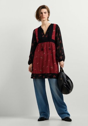 Frau, die ein rotes und schwarzes geblümtes Kleid über blauen Jeans trägt, hält eine schwarze Ledertasche und steht vor einem einfarbigen hellen Hintergrund.