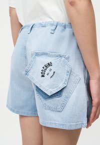 Ανοιχτό μπλε denim σορτς με δύο επικαλυπτόμενες πίσω τσέπες, μία τσέπη με την ετικέτα "Moschino 86-23 Jeans", φορεμένα με λευκή μπλούζα.