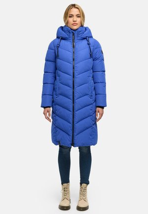 Vrouw draagt een lange blauwe gewatteerde winterjas met capuchon, donkere aangesloten jeans en beige veterlaarzen, staand tegen een effen achtergrond.