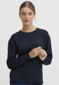 Mørkeblå hettegenser med rund hals, lange raglanermer og strukturert stoff. Har sømdetaljer langs fronten.
