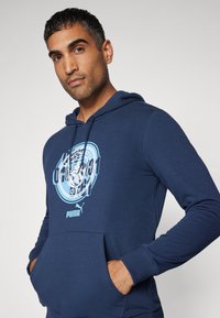 Felpa con cappuccio blu navy con tasca frontale, caratterizzata da un design grafico azzurro chiaro e dal logo Puma. Texture liscia e polsini aderenti.