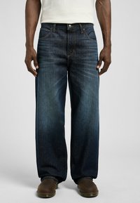 Dark blue denim jeans with a relaxed fit, featuring five-pocket design and contrasting stitching. Worn with brown footwear.