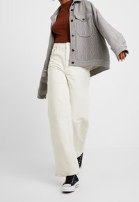 Personne portant un pantalon large en velours côtelé crème, des baskets montantes noires, un top marron ajusté et une veste à carreaux beige, en plein mouvement sur un fond uni.