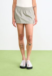 Osoba v svetlosivých mini sukni a strieborných slip-on topánkach, ktorá má tetovania na nohách a rukách, na jednofarebnom pozadí.