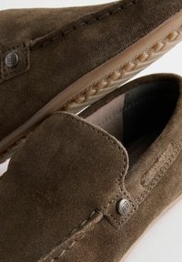 Braune Wildleder-Slipper mit gestickten Details und einem Metall-Logo-Akzent. Die Sohle ist flexibel und verfügt über ein strukturiertes Grip-Muster.