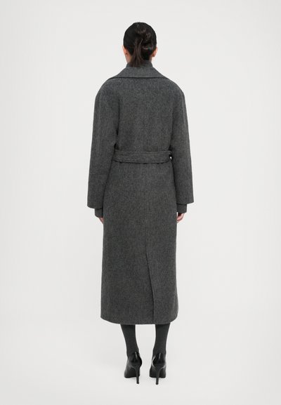 Long manteau en laine gris avec une taille ceinturée, un large col et une fente sur le côté. Présente un tissu texturé et une coupe droite, adapté à diverses tenues.