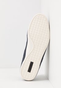 Granatowy sneaker z teksturowaną gumową podeszwą. Posiada białą, wzorzystą podeszwę zewnętrzną oraz czarną plakietkę z logo Lacoste. Górna część wykonana z gładkiej skóry.