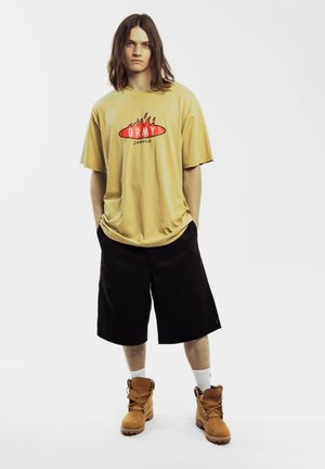 Gelbes, übergroßes T-Shirt mit rotem grafischem Design, kombiniert mit schwarzen knielangen Shorts und braunen Schnürstiefeln. Weiße Socken sind sichtbar.