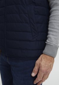 Jūrų karininkų puffy liemenė su quilted dizainu ir šonine kišene, derinama su šviesiai pilku raukšlėtu megztiniu ir tamsiai mėlynomis džinsinėmis kelnėmis.