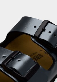 Κοντινή λήψη μαύρου δερμάτινου σανδαλιού με δύο ρυθμιζόμενους λουριά με αγκράφες και ταμπά πάτο ποδιού.